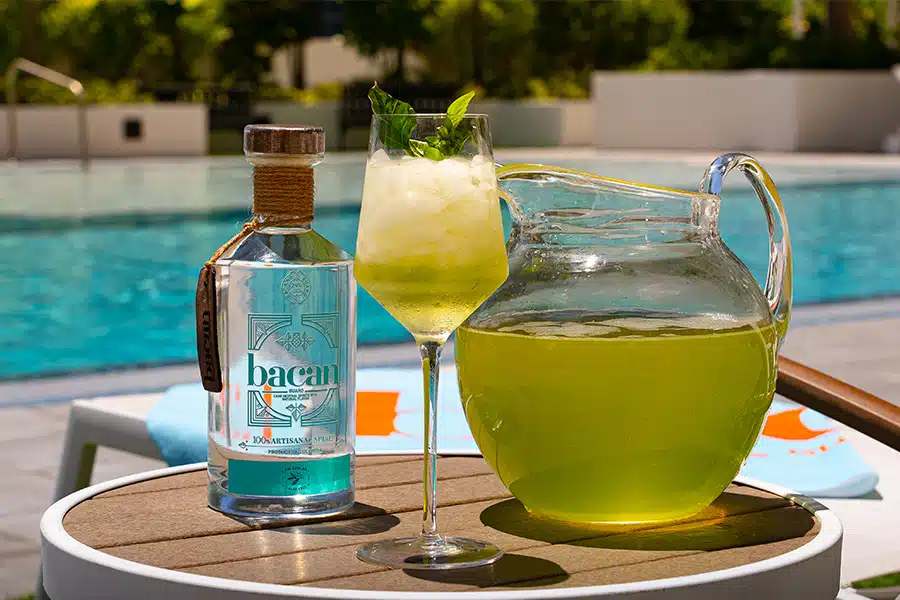 A bottle of Bacanora, a glass filled with a yellow-green iced drink garnished with mint, and a matching pitcher are on a table by a swimming pool on a sunny day.