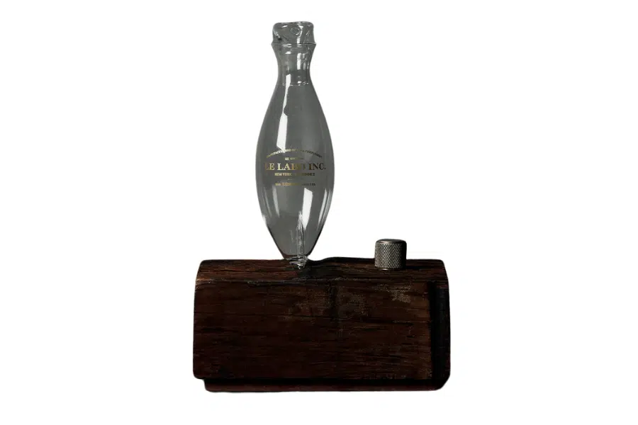 A clear glass bottle shaped like a fish is balanced upright on a dark wooden block. Next to it, there is a small metallic thimble resting on the block’s surface. The background is white.