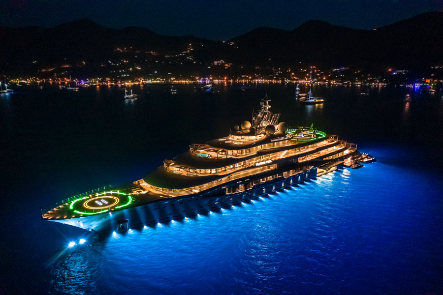 A large luxury yacht with vibrant lights is anchored at night in calm blue waters, with hills and twinkling city lights visible in the background.