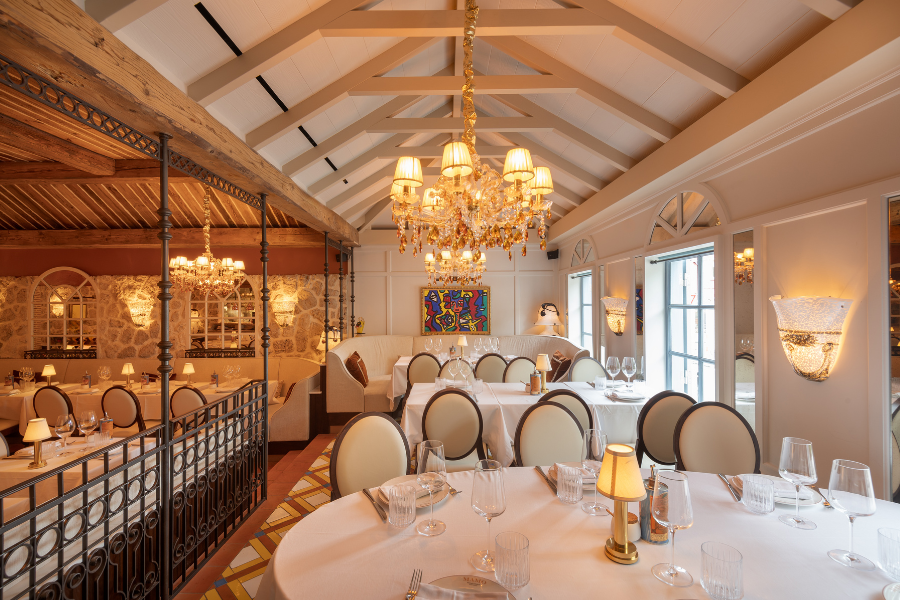 Elegant restaurant interior with round tables covered in white cloths, set with glassware. White cushioned chairs, chandeliers, large windows, and a colorful painting create a bright, upscale ambiance.
