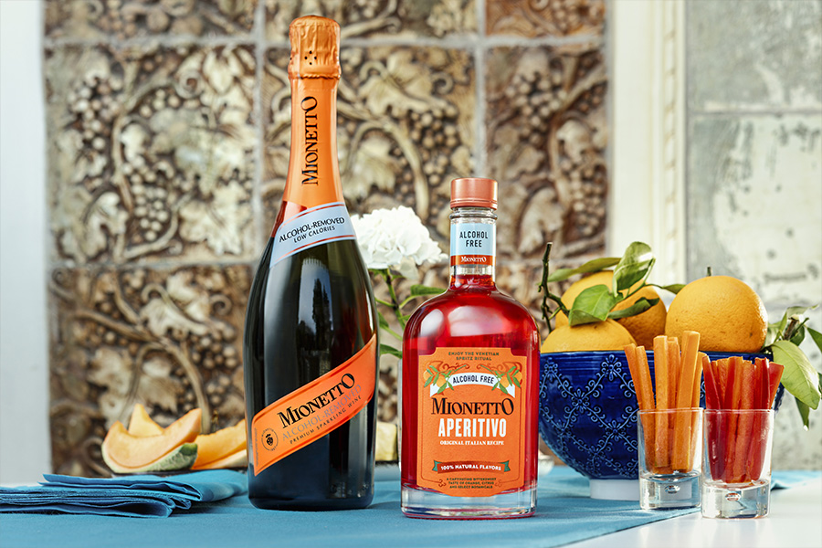 A bottle of Mionetto Prosecco and a bottle of Mionetto Aperitivo stand on a table with a blue cloth, surrounded by oranges, a blue bowl, cut fruit, and breadsticks, with a floral arrangement in the background.