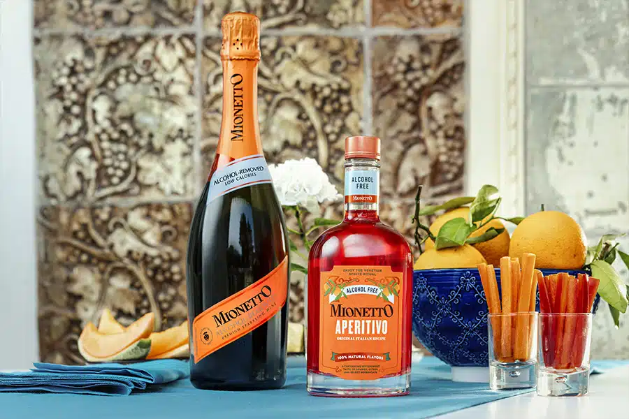 A bottle of Mionetto Prosecco and a bottle of Mionetto Aperitivo stand on a table with a blue cloth, surrounded by oranges, a blue bowl, cut fruit, and breadsticks, with a floral arrangement in the background.