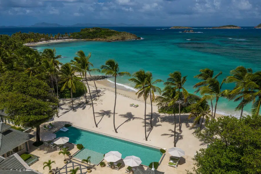 Aerial view of a tropical beach with turquoise water, white sand, tall palm trees, and a rectangular pool with lounge chairs and umbrellas. Lush greenery and distant islands are visible under a partly cloudy sky.