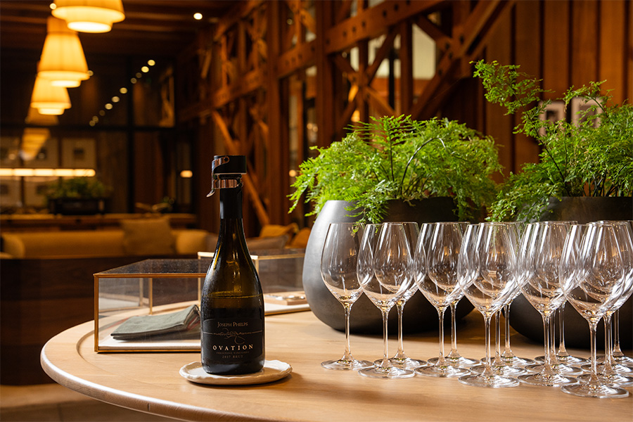 A bottle of sparkling wine and several empty wine glasses are arranged on a round wooden table, with green potted plants and warm, ambient lighting in an elegant, modern dining space.