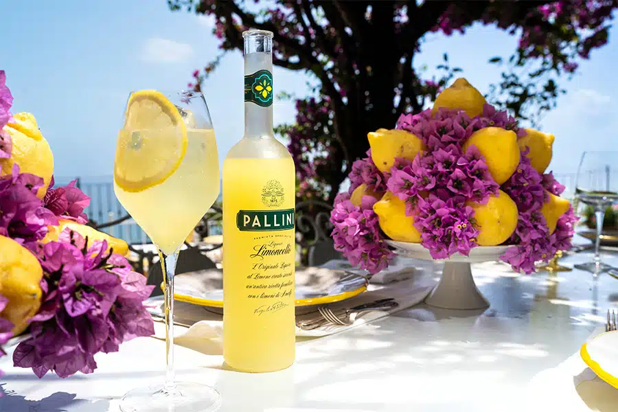 A bottle of Pallini Limoncello, a glass of lemon cocktail with a lemon slice, and a cake stand with lemons and purple flowers sit on an outdoor table in bright sunlight, with trees and blue sky in the background.