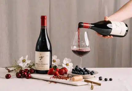 A hand pours red wine into a glass next to a bottle of wine, with fresh cherries, strawberries, blueberries, a corkscrew, cork, and white flowers arranged on a white table.