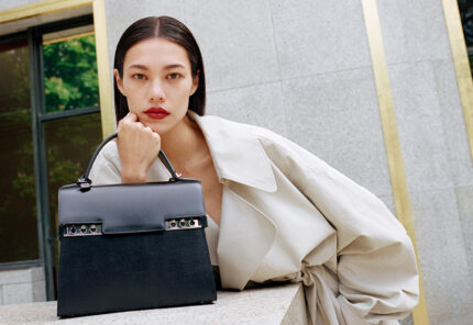 A woman with straight dark hair and red lipstick wears a light-colored trench coat and leans on a stone ledge, resting her face on her hand. A black handbag sits in front of her. The background features stone walls and windows.