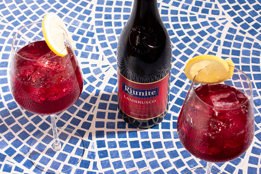 A bottle of Riunite Lambrusco is placed on a blue and white mosaic table, flanked by two wine glasses filled with red wine, ice, and garnished with lemon slices.