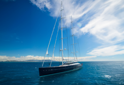 A large, modern sailing yacht with multiple masts glides on deep blue water under a bright, partly cloudy sky, with distant land visible on the horizon.