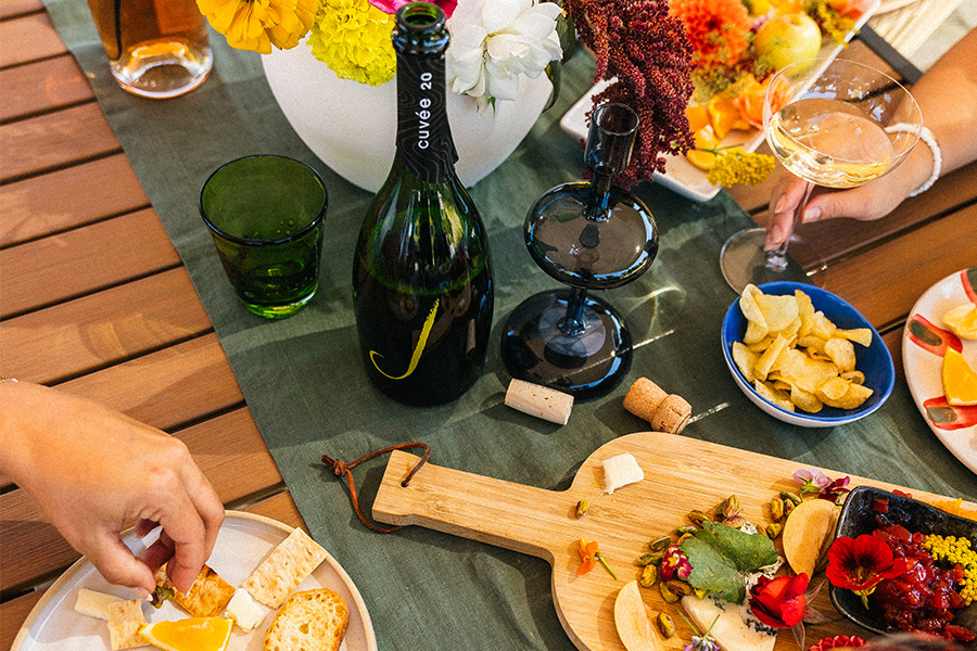 A colorful table setting with wine, cheese, crackers, chips, fruit, and flowers. Two people are reaching for food, and a bottle of wine and filled wine glasses are visible. The scene is bright and inviting.