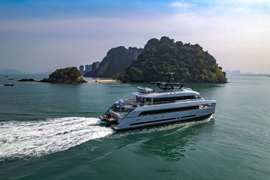 Southeast Asia yacht destinations A modern white yacht cruises on calm blue-green water near a small, lush island with rocky cliffs under a partly cloudy sky.
