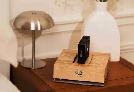 A wooden scent diffuser with a black cartridge sits on a wooden nightstand next to a modern silver lamp and a white vase.