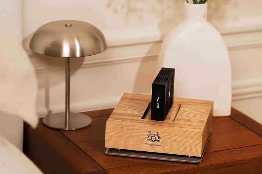 A wooden scent diffuser with a black cartridge sits on a wooden nightstand next to a modern silver lamp and a white vase.