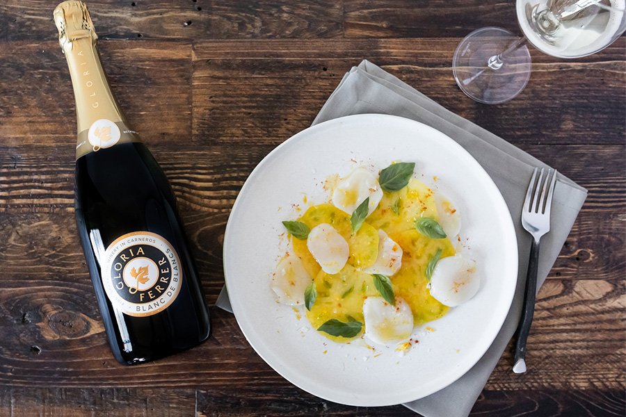 A plate of scallop carpaccio with herbs and olive oil sits next to a bottle of Gloria Ferrer sparkling wine on a wooden table, with a wine glass, fork, and gray napkin nearby.