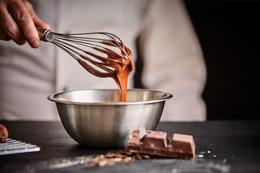 A person whisking melted chocolate in a metal bowl, with chocolate blocks and shavings on a dark surface in the foreground.