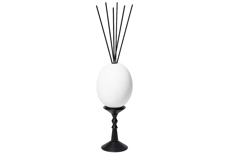A white oval-shaped diffuser with black reed sticks is placed on a black, decorative stand against a plain white background.