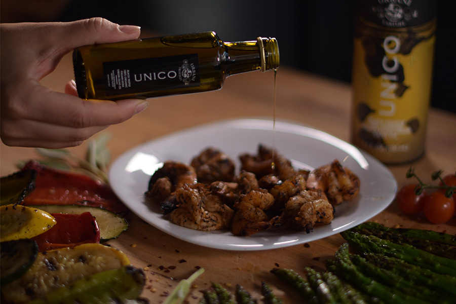 A hand pours olive oil from a bottle labeled UNICO onto a plate of grilled shrimp. Roasted vegetables, asparagus, and cherry tomatoes surround the plate on a wooden table.