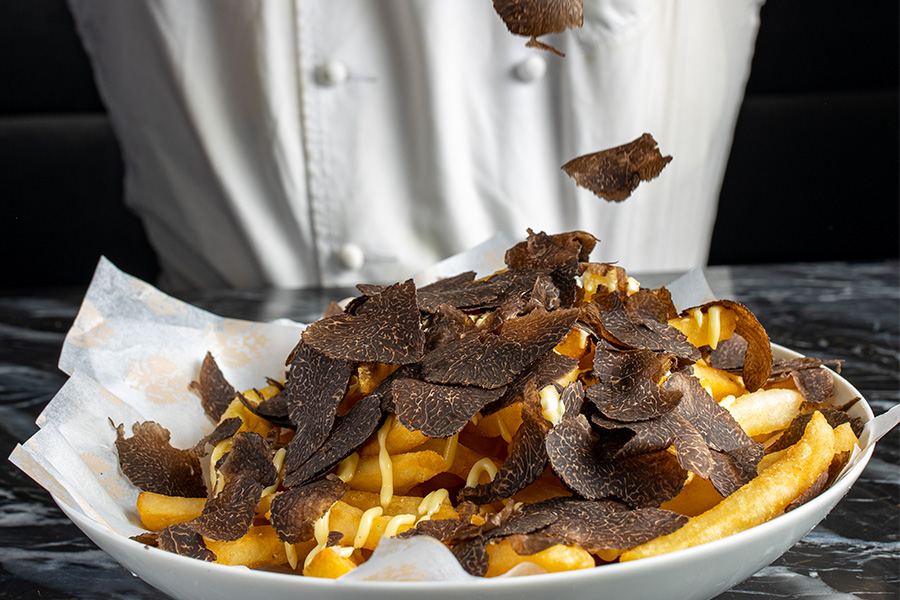 A chef sprinkles generous shavings of black truffle over a plate of golden French fries drizzled with sauce, served on parchment paper atop a dark marble table.