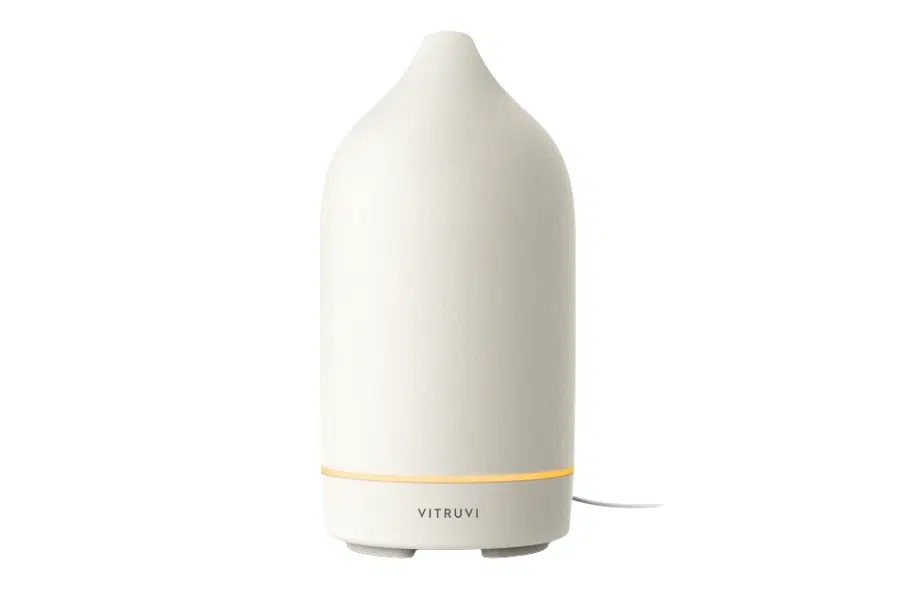 A minimalist, off-white ceramic essential oil diffuser with a soft amber light at the base, labeled Vitruvi, sits on a white background.
