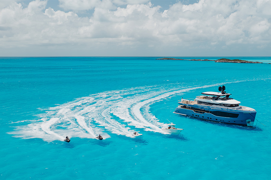 A large blue yacht is anchored on bright turquoise water, surrounded by four jet skis creating white trails as they speed nearby under a partly cloudy sky.