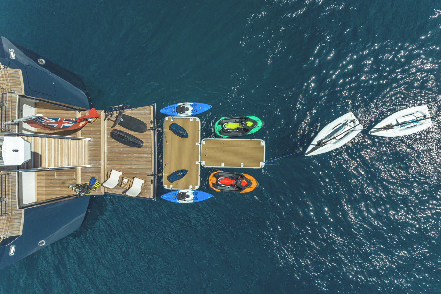 Aerial view of a yacht with a wooden deck, lounge chairs, and water toys including jet skis, kayaks, and two small sailboats floating on calm blue water.