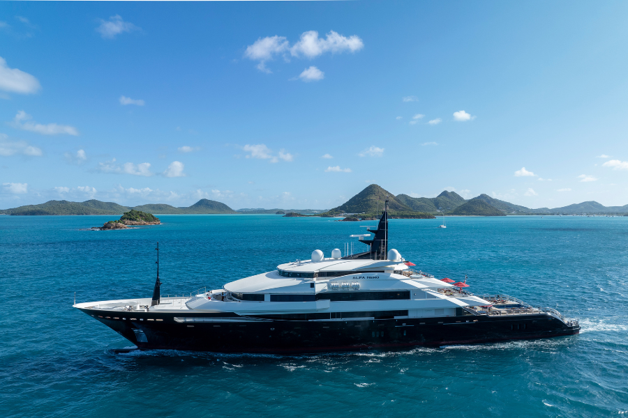 A large luxury yacht sails on clear blue water with small islands and green hills in the background under a bright blue sky with scattered clouds.