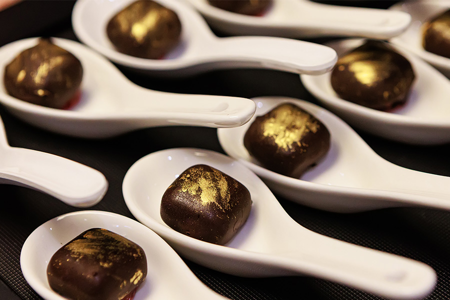 Several white ceramic spoons, each holding a small square of dark chocolate decorated with gold accents, arranged neatly on a black surface.