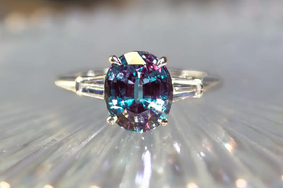 A close-up of a silver ring with a round, multicolored gemstone, displaying shades of blue, purple, and teal, set on a blurred, reflective surface.