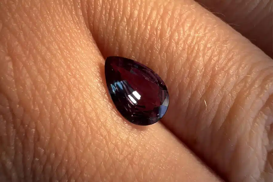 A close-up of a dark red pear-shaped gemstone resting on the textured skin of a persons hand, reflecting light on its polished surface.