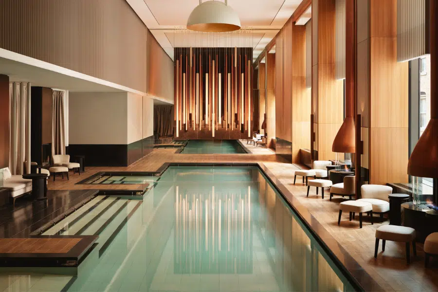 A luxurious indoor pool area with modern wooden accents, high ceilings, elegant pendant lighting, comfortable lounge chairs, and large windows allowing natural light to fill the sophisticated space.
