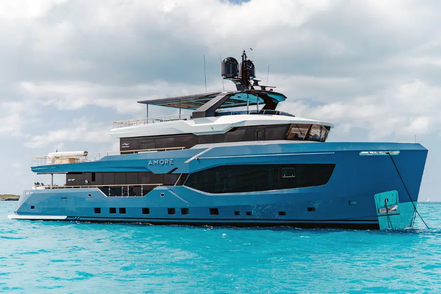 A modern blue luxury yacht named AMORE is anchored on clear turquoise water under a partly cloudy sky.
