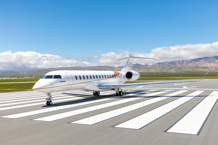 A sleek white private jet with gold accents is parked on a runway at an airport, surrounded by green fields and distant mountains under a partly cloudy sky.