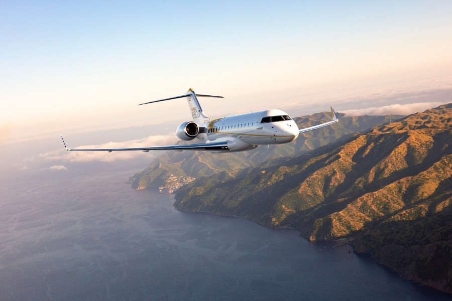 A white private jet flies over a coastal mountain range with the ocean below, under a clear sky during daylight.