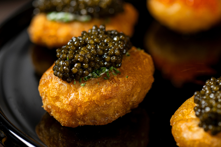 A close-up of a golden, crispy fried appetizer topped with a generous portion of black caviar and a hint of green garnish, served on a glossy black plate.