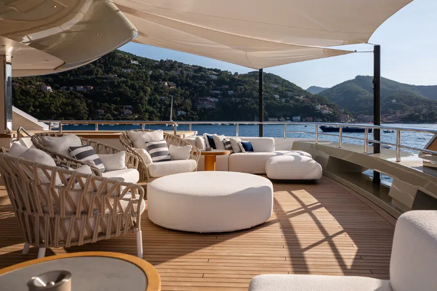 Luxurious yacht deck with cushioned lounge chairs, a round ottoman, and sunshades. The deck overlooks calm blue water and green, hilly coastline under a clear sky.