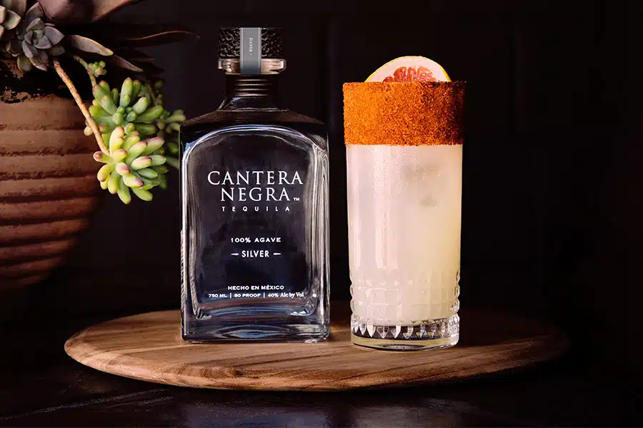 A bottle of Cantera Negra Silver Tequila sits next to a tall glass of a light cocktail with a chili-rimmed glass and grapefruit slice garnish, on a wooden tray beside a potted succulent.