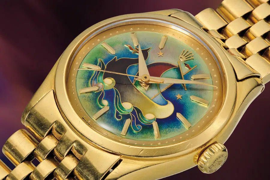 A gold wristwatch with a colorful, abstract dial featuring whimsical shapes, stars, and swirls. The watch has a gold metal strap and a textured crown, set against a blurred purple background.