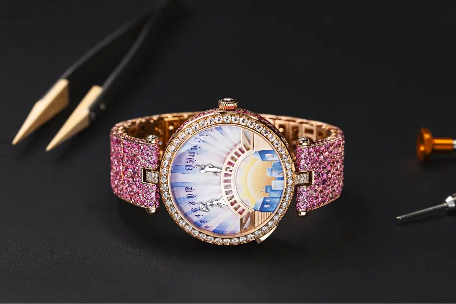 A luxurious watch with a pink jeweled band and a decorative face featuring a sun, blue sky, and castle-like buildings, displayed on a black surface with watchmaking tools nearby.