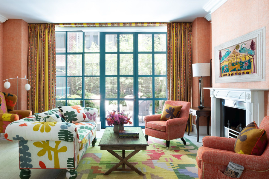 Colorful living room with a floral-print sofa, two orange armchairs, a wooden coffee table, patterned rug, large window with yellow curtains, wall art above a white fireplace, and fresh flowers on the table.
