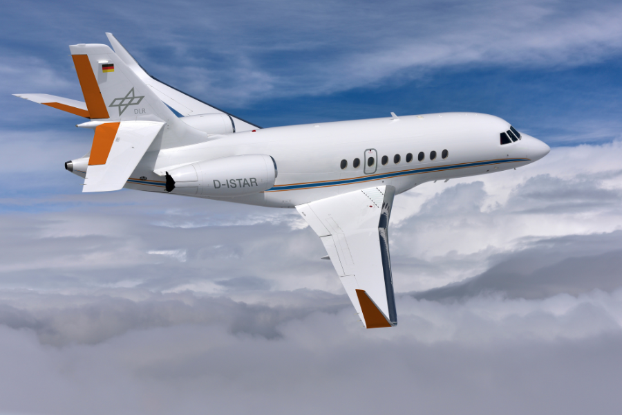 A white Dassault Falcon jet with orange accents and the German Aerospace Center logo flies above clouds against a blue sky. The aircraft registration reads D-ISTAR.