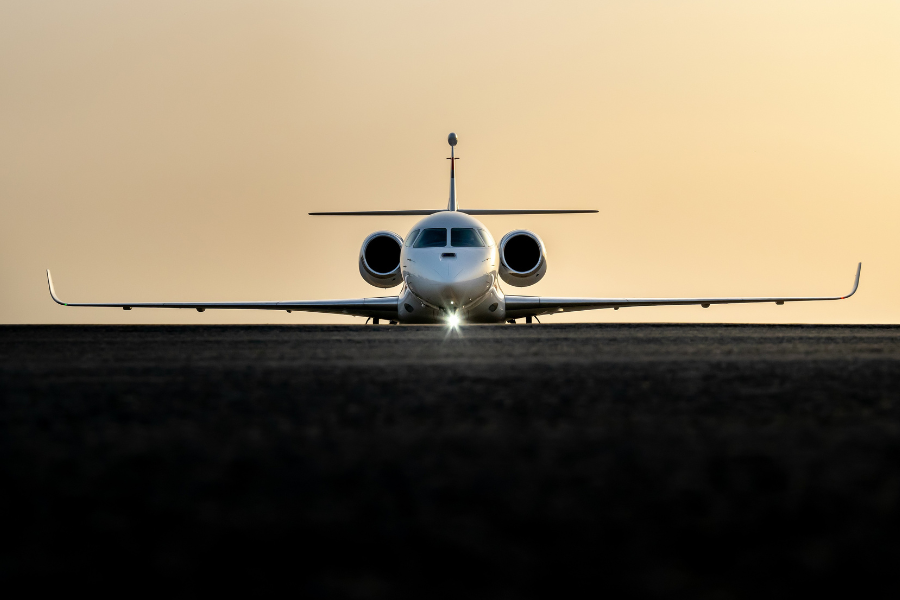 A private jet photographed from the front sits on a runway at sunset, with its lights on and wings outstretched, against a softly glowing sky.