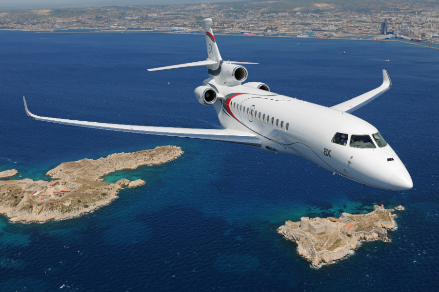 A sleek white private jet with red and grey accents flies over deep blue coastal waters and small rocky islands, with a cityscape and hills visible in the distant background.