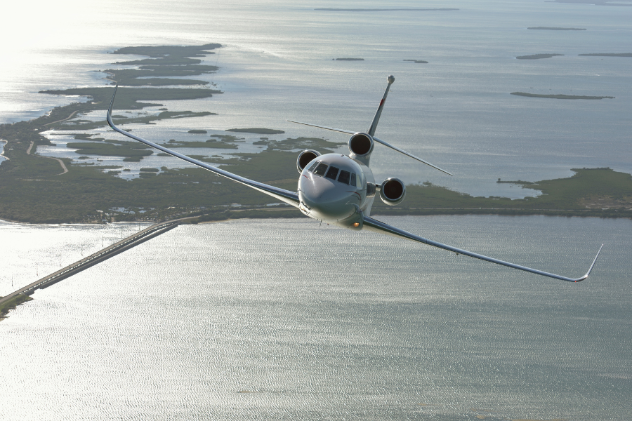 A private jet flies low over a large body of water, with islands and a bridge visible below on a sunny day. The aircraft is captured head-on, banking slightly with its wings angled.