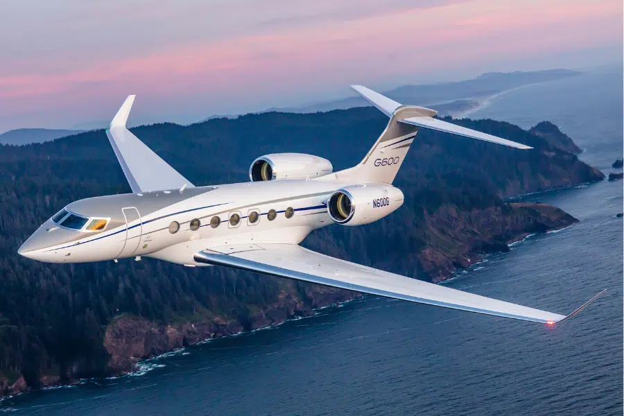 A white and silver private jet flies over a rugged coastline with dense forests at sunset, with the ocean and distant hills visible beneath a colorful sky.
