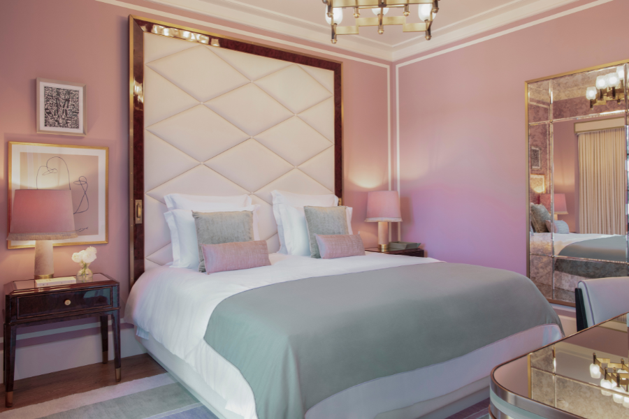 A stylish bedroom with pastel pink walls, a large padded headboard, a neatly made bed with white and light blue bedding, bedside tables with lamps, and modern artwork on the walls.