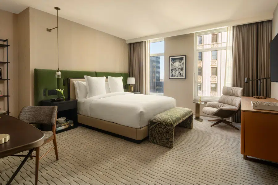 A modern hotel room with a large bed, green headboard, and white bedding, a patterned bench at the foot, an armchair by the window, a desk, and beige walls with large windows letting in natural light.