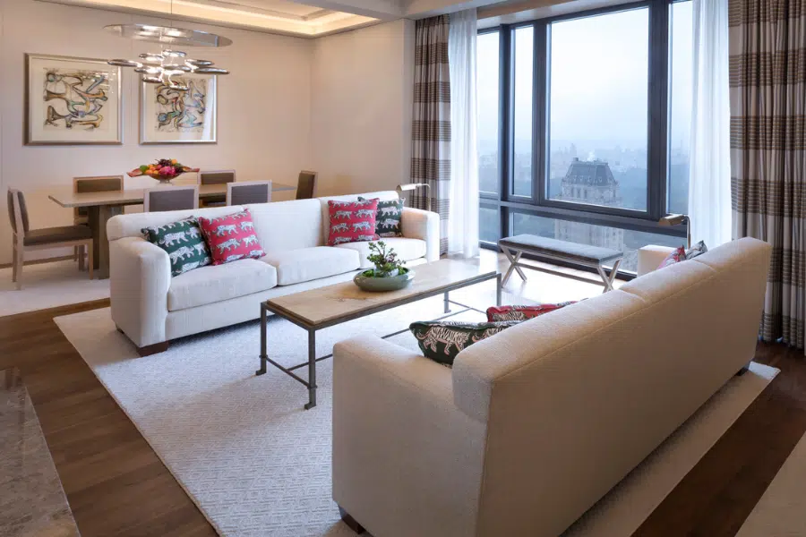 Modern living room with two white sofas, colorful patterned pillows, a coffee table, large windows with city views, and a dining area with wooden furniture and contemporary art on the walls. Neutral tones with pops of color.