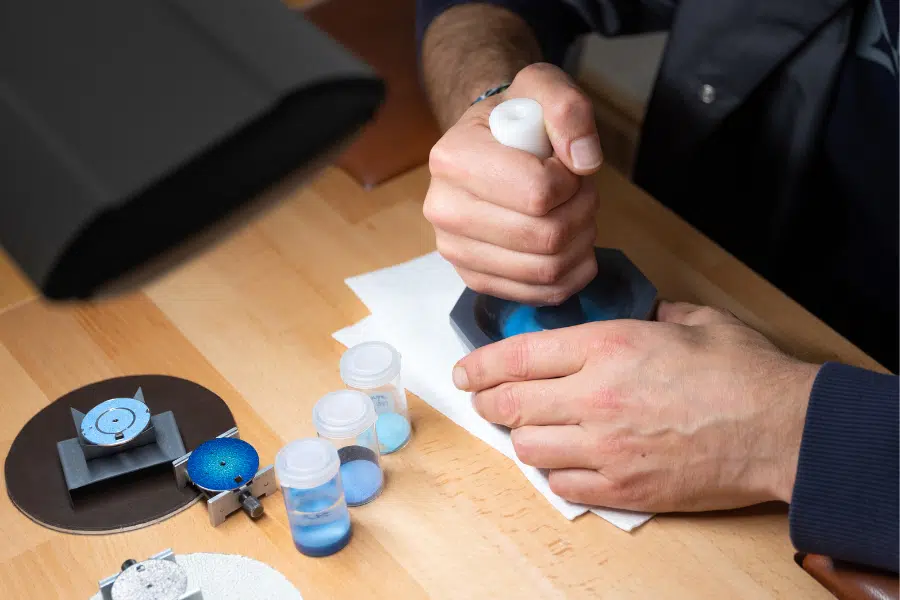 A person uses a tool to polish or grind a small blue object on a workbench. Nearby are jars of blue powder, a round metal object, and other watchmaking or crafting tools.