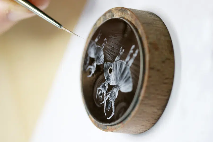 A close-up of a hand painting two white elephants on a small, round, dark surface with a thin brush. The artwork is detailed and enclosed in a wooden frame.