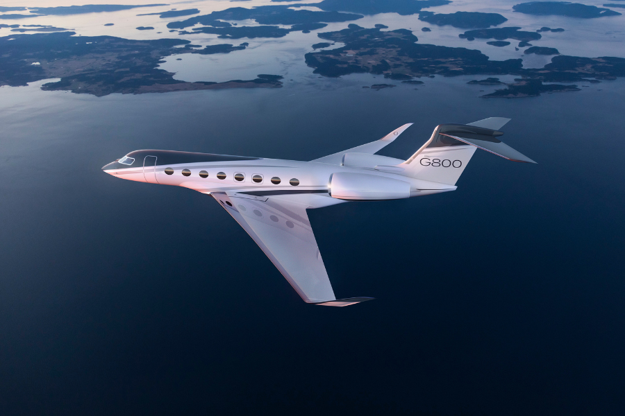 A sleek white Gulfstream G800 private jet flies over a body of water with scattered islands below, reflecting the soft light of sunset or sunrise.